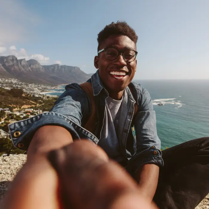 Homem jovem negro sorridente, usando óculos e camisa jeans, sentado em um mirante natural à beira-mar. Ele estende a mão em direção à câmera em um gesto de convite. Ao fundo, uma costa montanhosa e o oceano sob um céu azul.