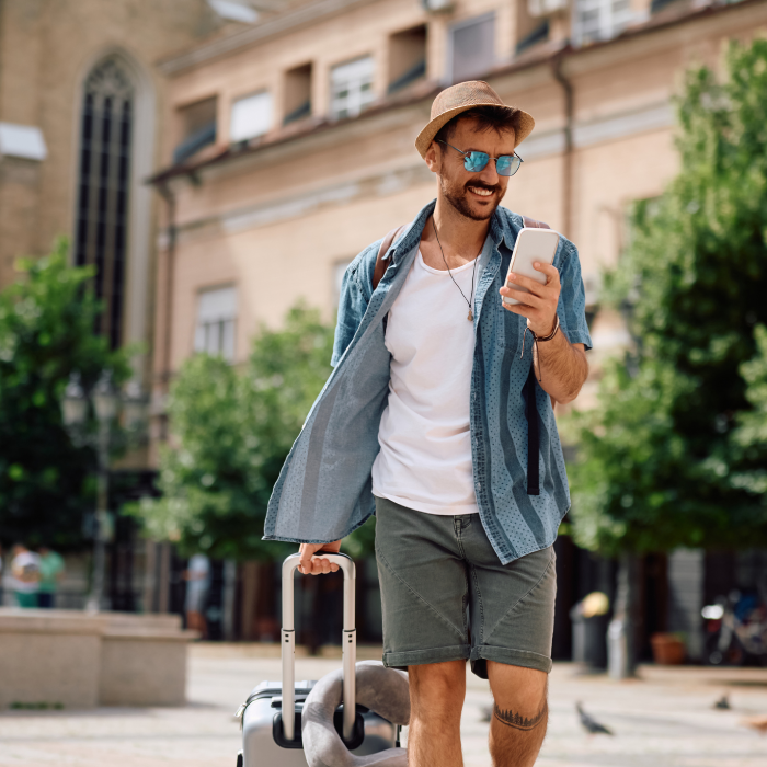 Homem caucasiano em estilo casual de viagem caminha por centro histórico urbano. Ele sorri enquanto usa o celular, carregando uma mala de mão e vestindo bermuda cinza e chapéu.
