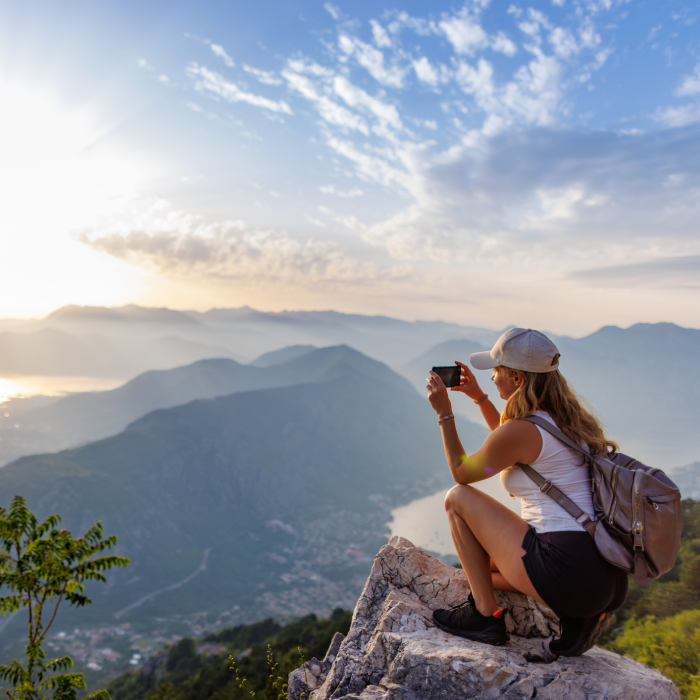 Uma mulher loira, vista de perfil, está agachada no topo de uma rocha em uma montanha elevada. Ela usa boné bege, regata branca, shorts pretos e uma mochila cinza, enquanto segura um smartphone para fotografar a paisagem. À sua frente, estende-se um vasto vale com montanhas cobertas por uma névoa suave, um rio ou baía sinuosa e uma cidade ao fundo. O sol está se pondo ou nascendo no lado direito, criando um brilho intenso no céu azul com poucas nuvens e refletindo na água lá embaixo.